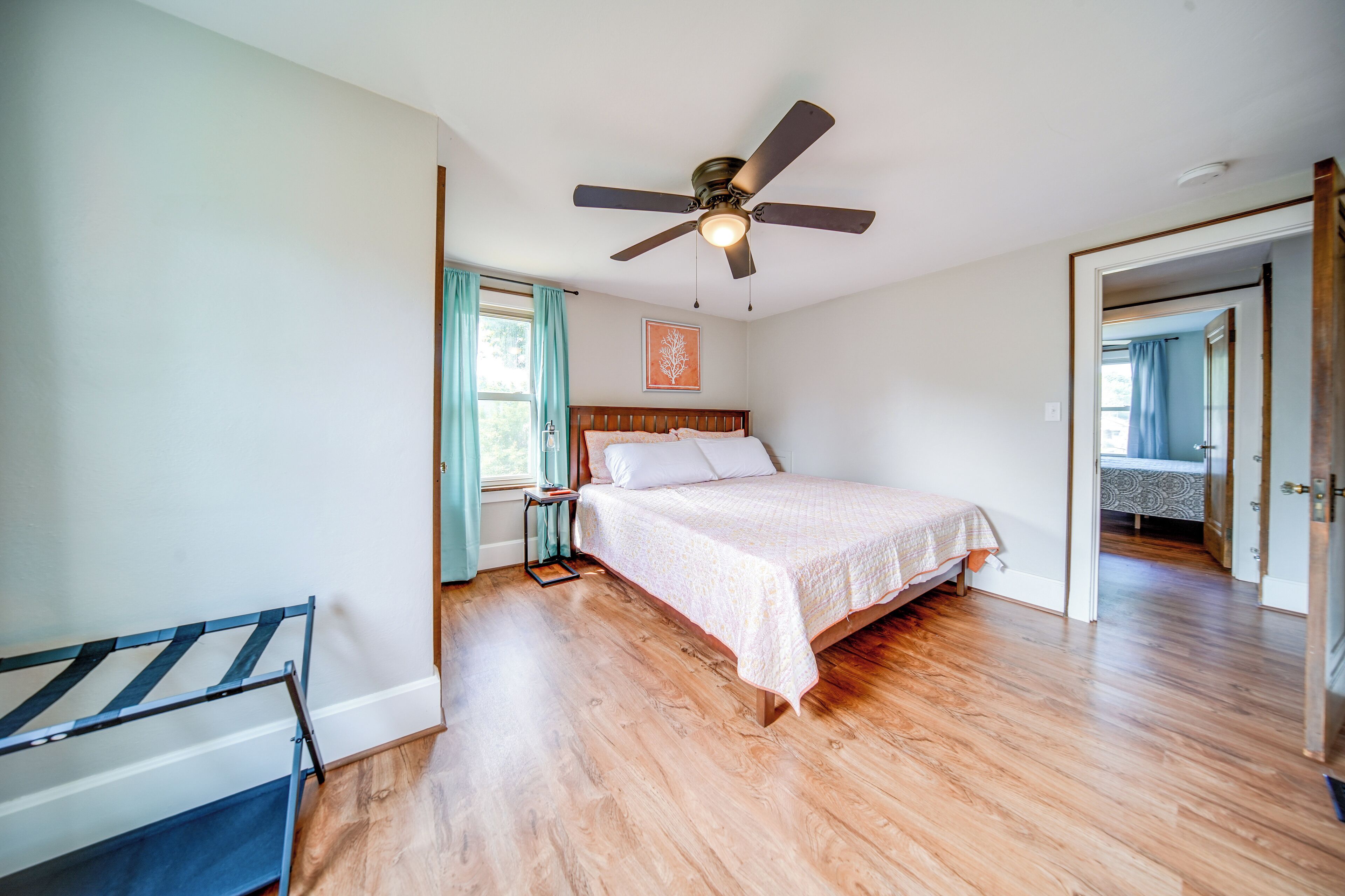 A bedroom with a bed, wooden flooring, a ceiling fan, a window with curtains, and a door leading to another room.