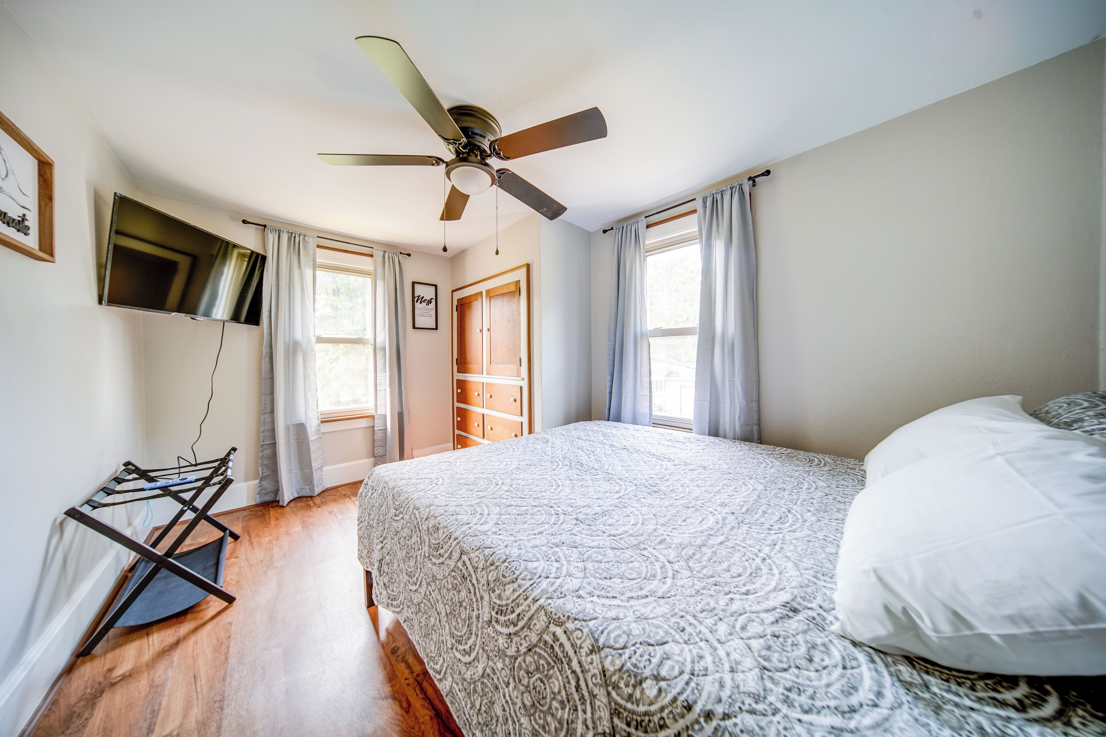 A bedroom with a bed, a ceiling fan, a TV mounted on the wall, and a window with curtains.