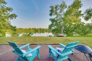 Two turquoise Adirondack chairs facing a calm lake with a grassy area and trees in the background.