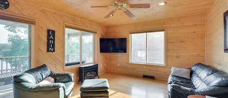 A cabin interior with wooden walls, a ceiling fan, a TV, leather sofas, and a fireplace.
