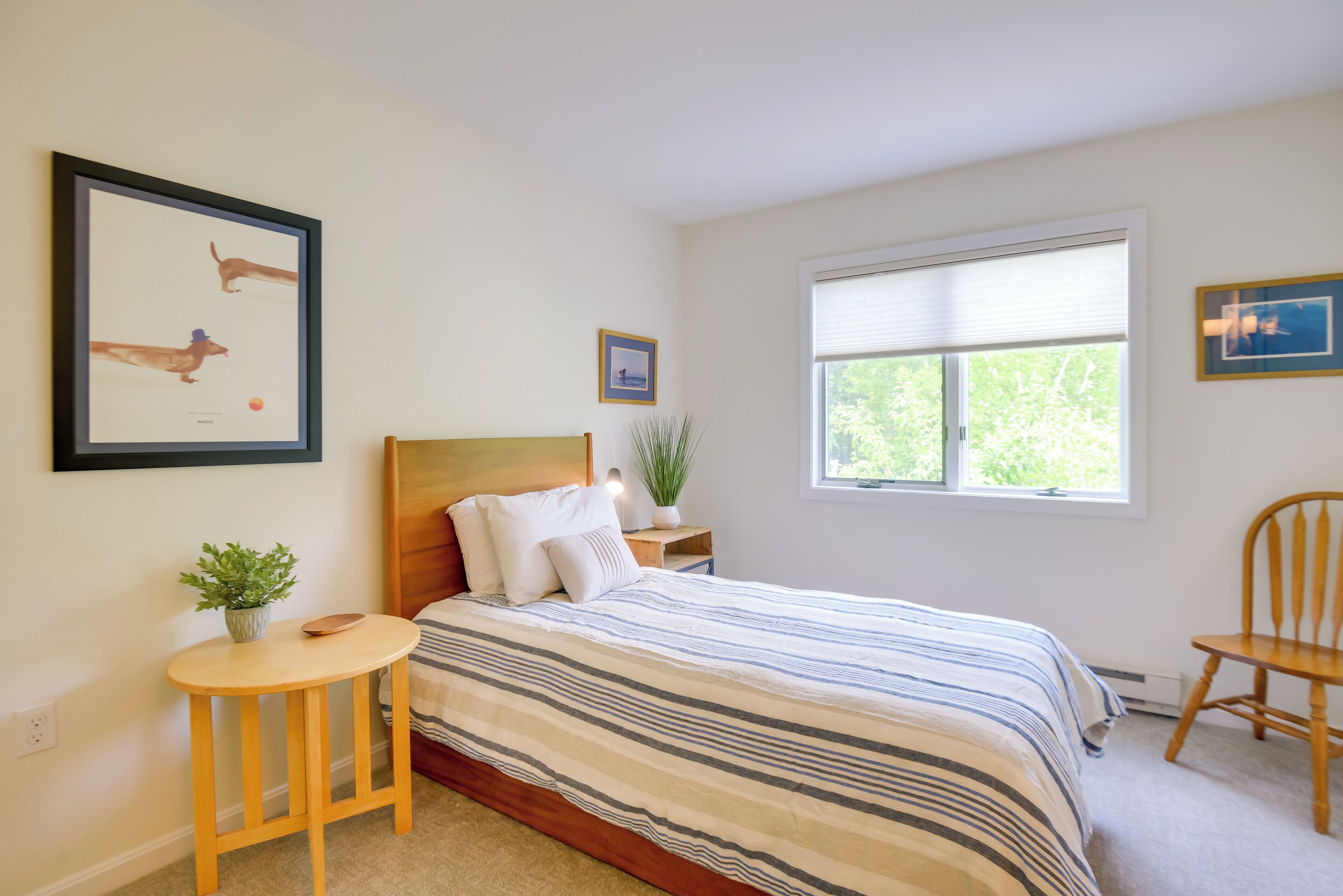A bedroom with a bed, a small table with a plant, a chair, and framed pictures on the wall.