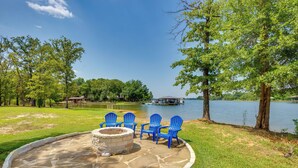 A stone fire pit with four blue Adirondack chairs around it, set in a grassy area with a lake and trees in the background. - On Cedar Creek Reservoir: Malakoff Home w/ Pool! (Malakoff)