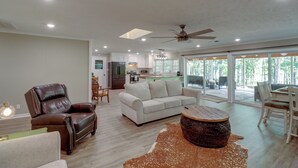 A spacious living room with a leather armchair, a sofa, and a coffee table. There is a kitchen in the background with a dining area visible through large windows. - Arkansas Vacation Rental on Greers Ferry Lake! (Higden)