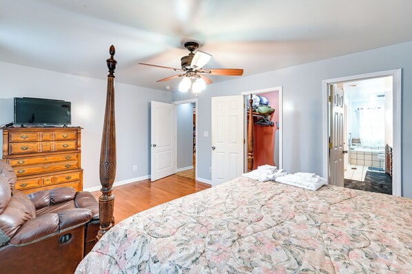 Une chambre à coucher comprenant un lit, une commode, un téléviseur, un ventilateur de plafond et une porte donnant sur une salle de bain.