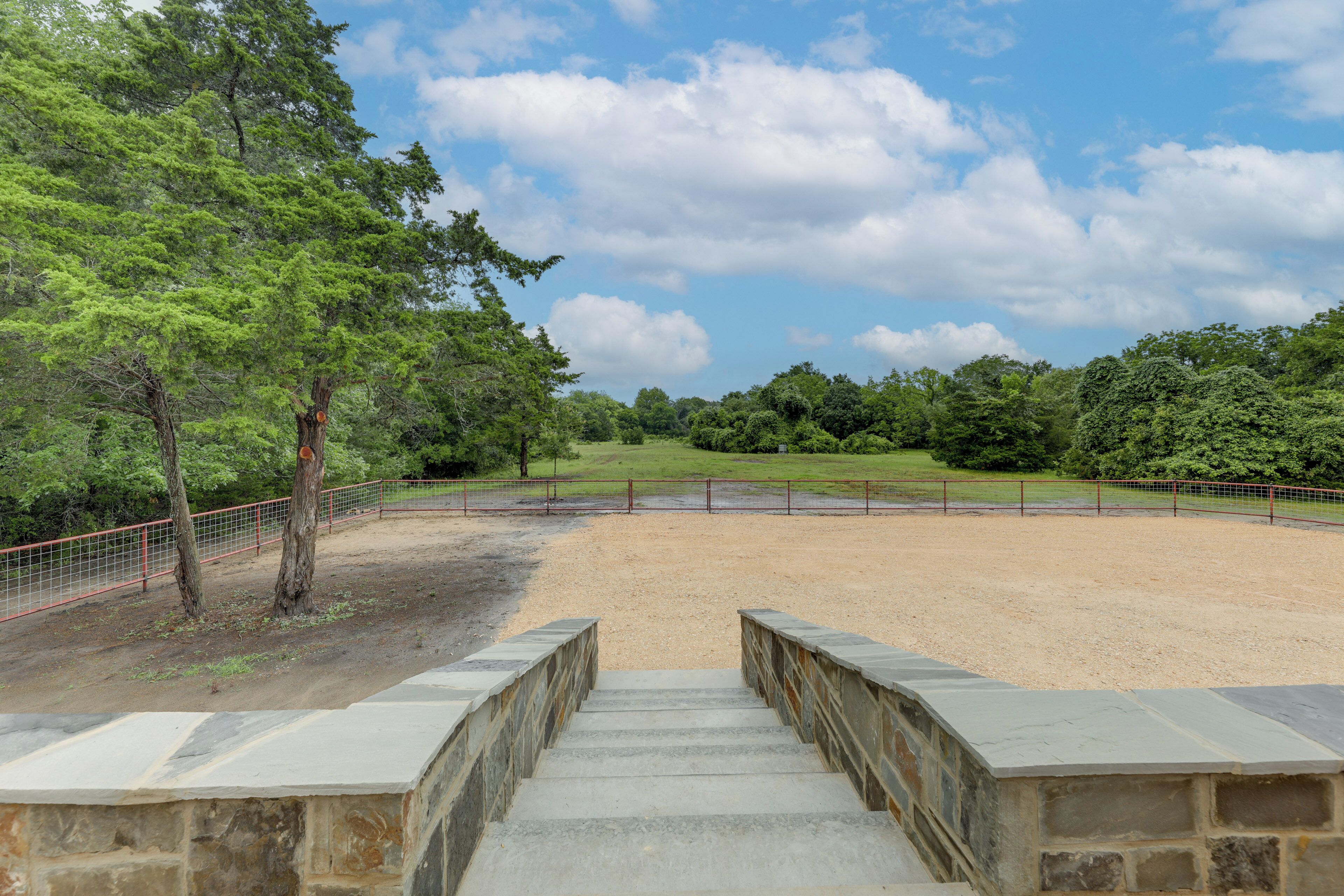 A stone staircase leading to a fenced arena with a dirt ground, surrounded by trees and greenery under a partly cloudy sky.
