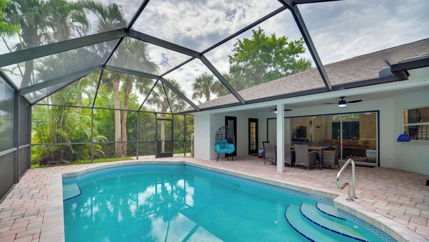 A swimming pool with a glass enclosure, surrounded by a patio area with outdoor furniture and a view of palm trees. - 'casa Blanco' Escape w/ Fire Pit in Sebastian! (Sebastian)