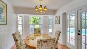 A dining area with a round table, four chairs with vintage patterns, a chandelier, and large windows with a view of greenery and a swimming pool. - 'casa Blanco' Escape w/ Fire Pit in Sebastian! (Sebastian)