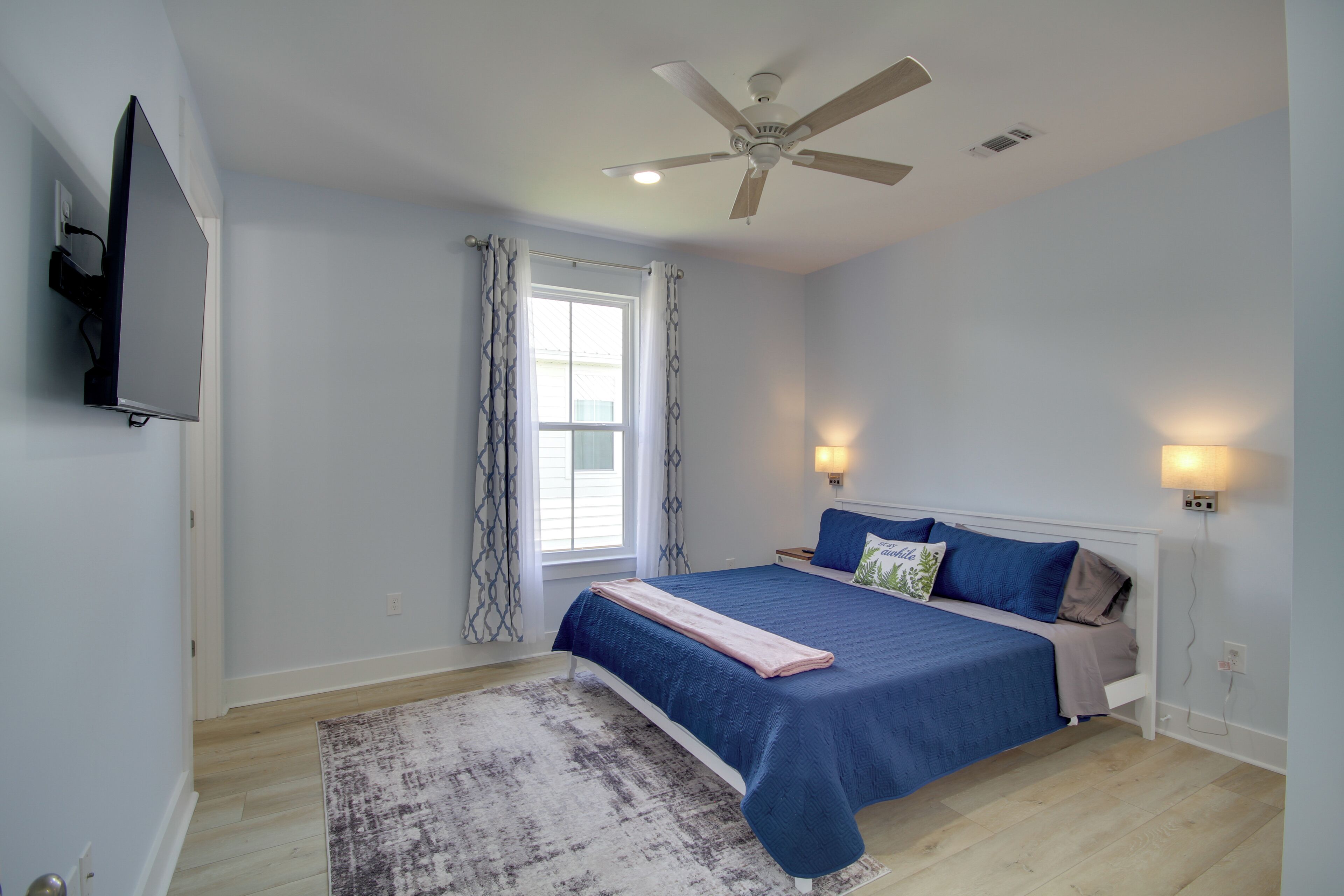 A bedroom with a bed, ceiling fan, window with curtains, wall-mounted TV, bedside lamps, and a rug.