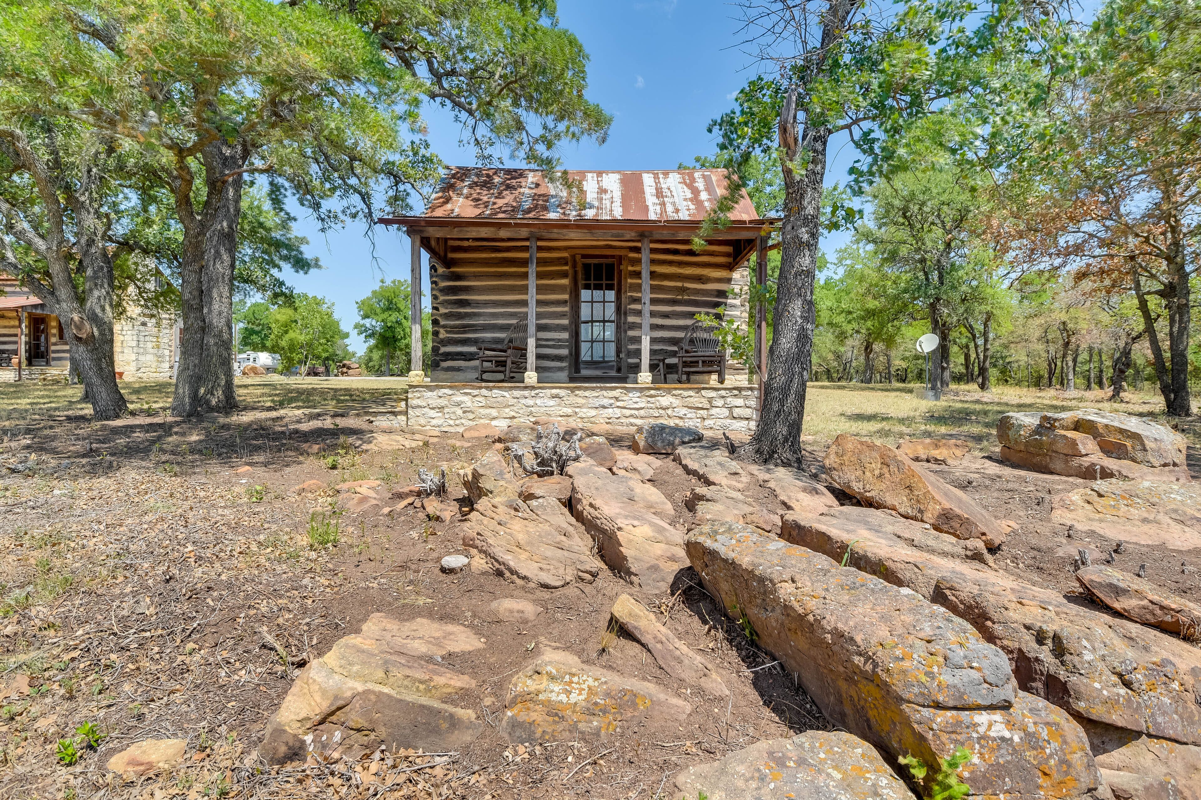 Une cabane rustique en rondins de bois, posée sur des fondations en pierre, et entourée de gros rochers et d’arbres.
