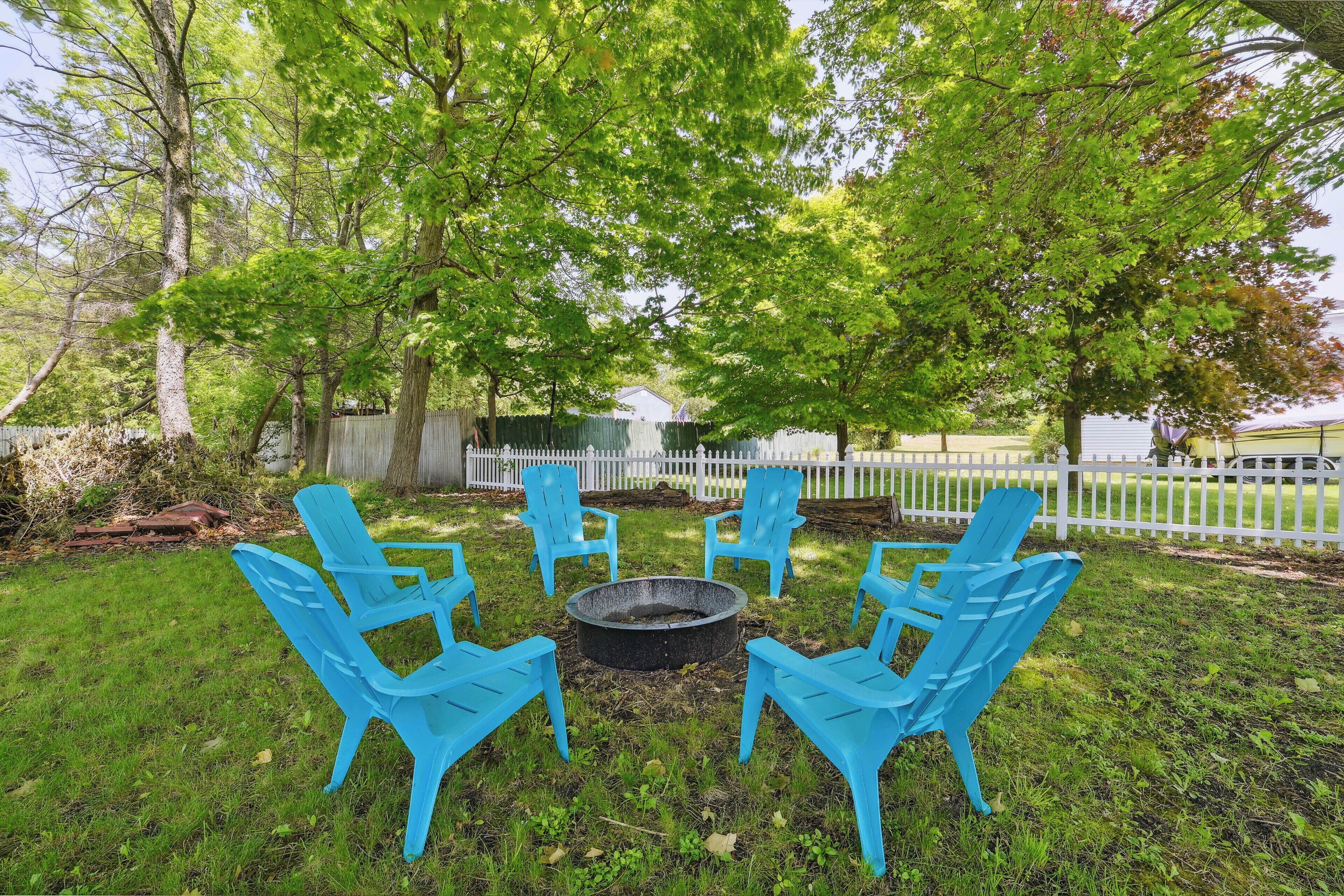 A backyard with a fire pit surrounded by four blue chairs, a white fence, and trees.