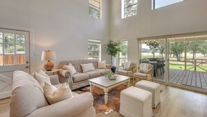 Un salon spacieux avec un haut plafond, de grandes fenêtres, une terrasse en bois et un coin salon confortable.