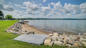 A lakeside view with a rocky shoreline, clear water, and a distant cityscape. - Luxury Lakefront Boyne City Townhome w/ Views! (Boyne City)