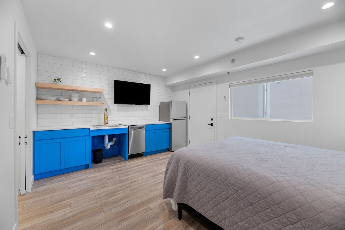 A modern bedroom with a large bed, a built-in wardrobe, and a small kitchenette with blue cabinets and a white tiled backsplash.