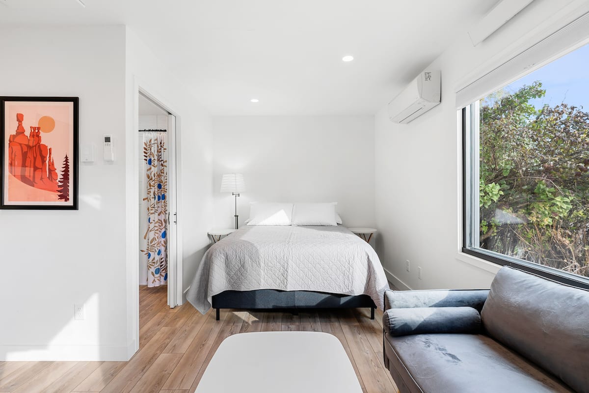 A modern bedroom with a bed, a sofa, a window with a view, and a door leading to another room.