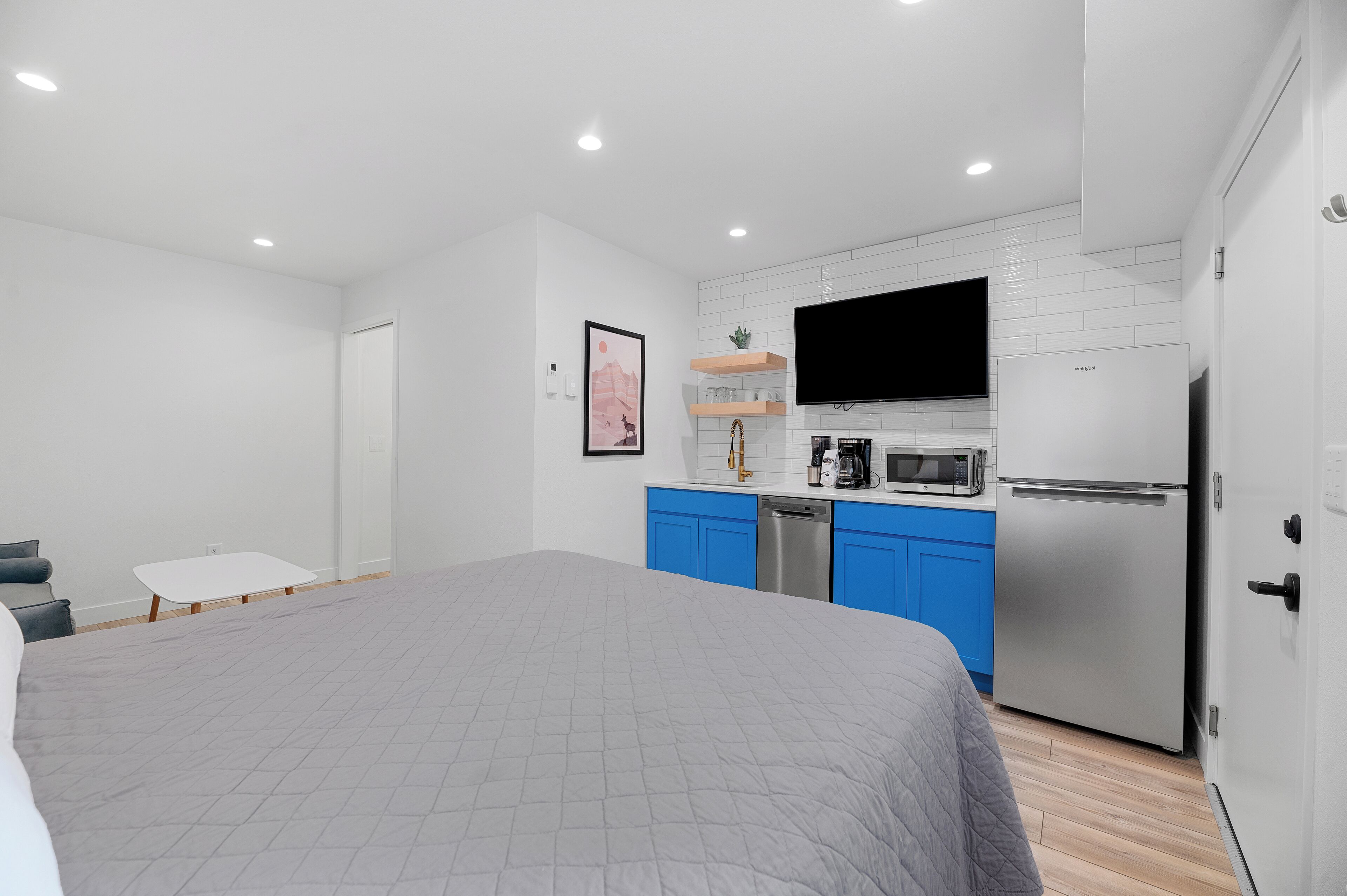 A bedroom with a bed, a small table, and a kitchenette with a microwave, toaster, and refrigerator.