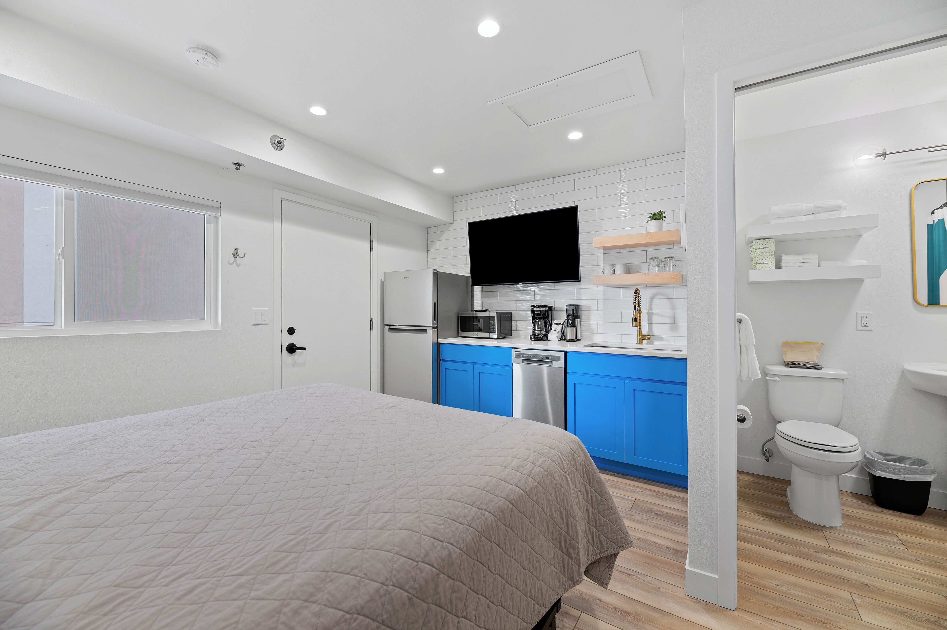 A modern bedroom with a large bed, a kitchenette with blue cabinets, a flat-screen TV, and a white toilet.