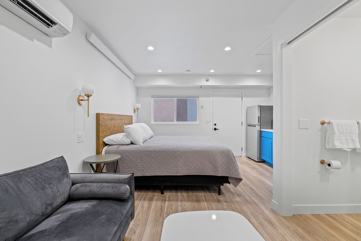 A modern bedroom with a bed, a grey sofa, a small round table, and a white bedside table.