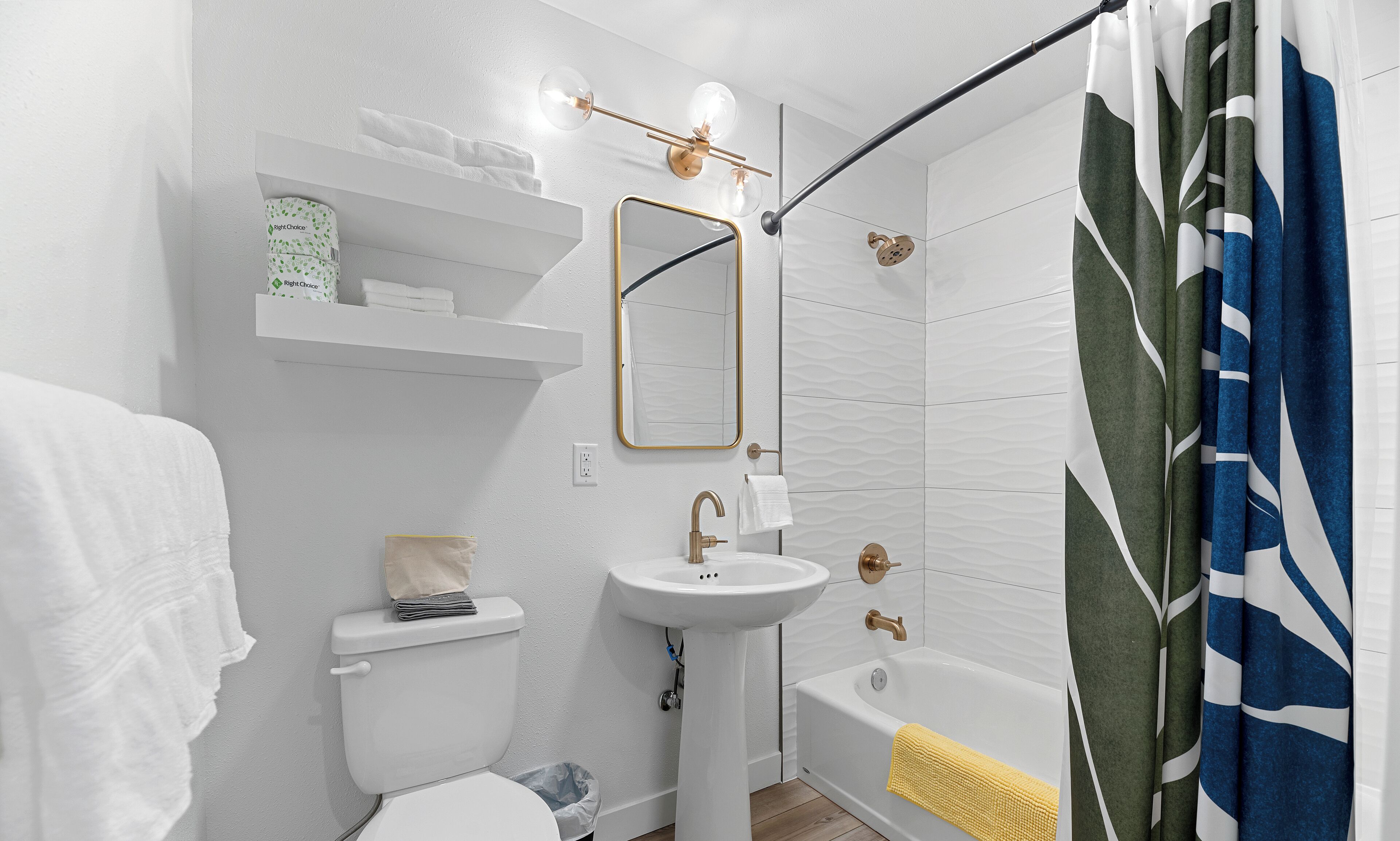 A modern bathroom with a white toilet, pedestal sink, and bathtub. There is a towel rack with folded towels, a mirror above the sink, and a shower curtain with a leaf pattern.