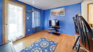A room with blue walls, a wooden floor, a TV on a stand, a patterned rug, and a dining table with chairs. - Pet-friendly Lake Erie Cottage - Walk to the Water (Geneva)