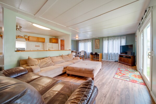A spacious living room with a sectional sofa, a coffee table, and a television. There is a kitchen area with wooden cabinets and a dining area with a table and chairs.