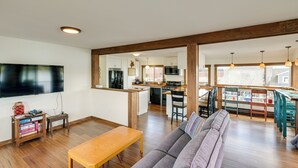 A spacious living room with a wooden bench, a gray sofa, and a TV mounted on the wall. A kitchen with modern appliances and a dining area with chairs and a table are visible in the background. - Ocean-view Apt in the Heart of Southwest Harbor! (Southwest Harbor)