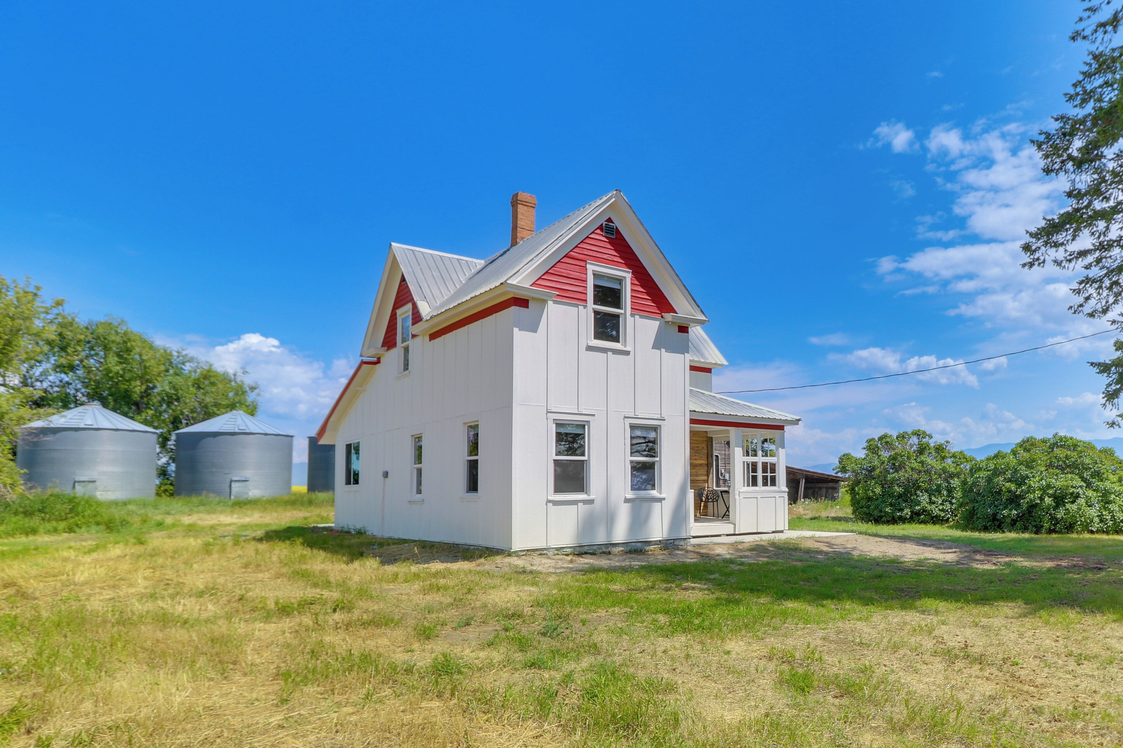 A two-story white house with a red roof and a chimney, surrounded by a grassy field and two large silos.