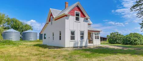 A two-story white house with a red roof and a chimney, surrounded by a grassy field and two large silos.