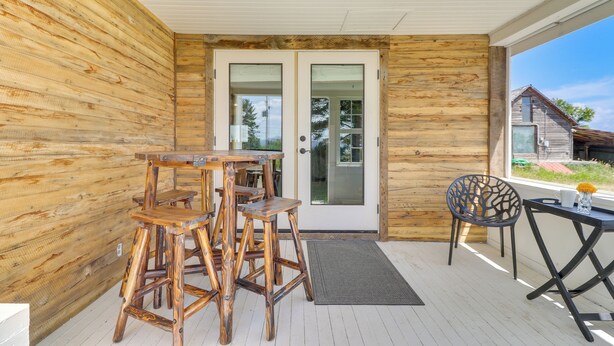 Un mur extérieur en bois, une porte en verre, une table en bois avec des chaises, une chaise noire et une petite table avec un vase et des fleurs.