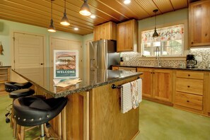 A kitchen with wooden cabinets, a stainless steel refrigerator, a coffee maker, and a welcome sign. - Unique Cotter Getaway w/ Deck & Outdoor Dining! (Cotter)