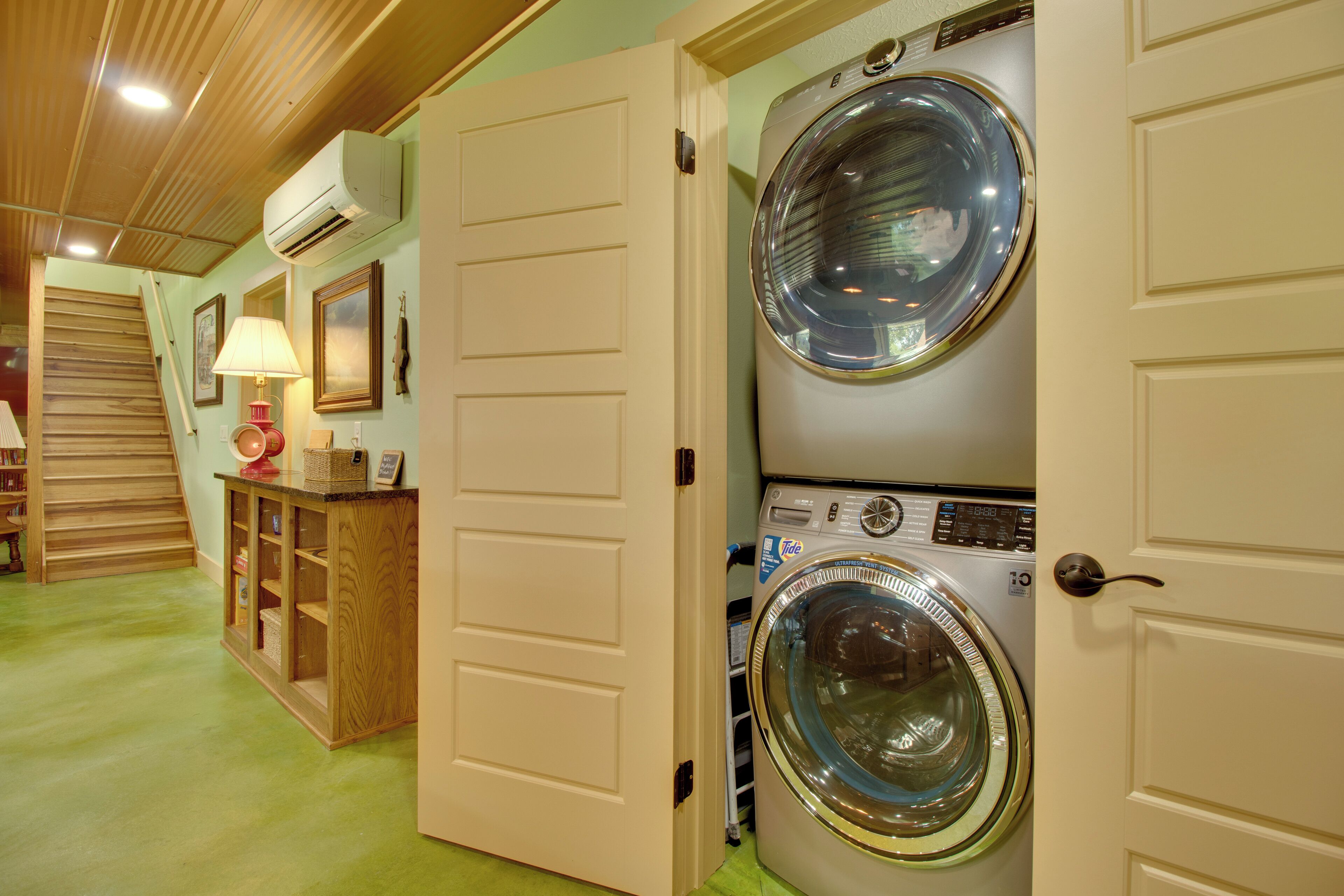 A laundry room with two stacked front-loading washing machines, a wooden cabinet with baskets, and a lamp on a shelf.