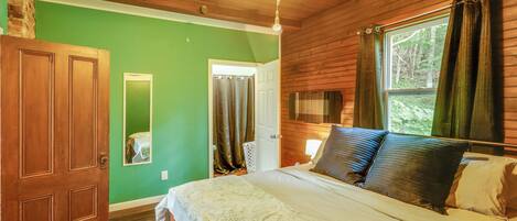 A bedroom with a wooden ceiling, a bed with patterned bedding, a nightstand, a mirror, and a window with curtains.