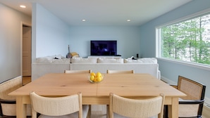 A modern dining area with a wooden table and wicker chairs, a bowl of fruit on the table, a sofa in the background, and a TV mounted on the wall. - Nisswa Lake Getaway w/ Hot Tub + Kayaks! (Nisswa)
