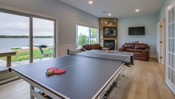 A ping pong table in a room with a stone fireplace, leather sofas, and a view of a lake. - Nisswa Lake Getaway w/ Hot Tub + Kayaks! (Nisswa)