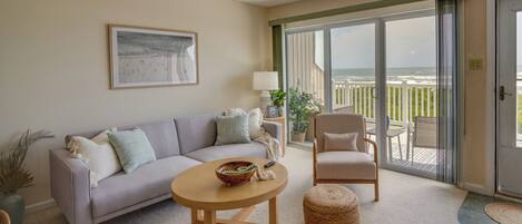 A living room with a sofa, armchair, round coffee table, and a view of the ocean through large sliding glass doors.