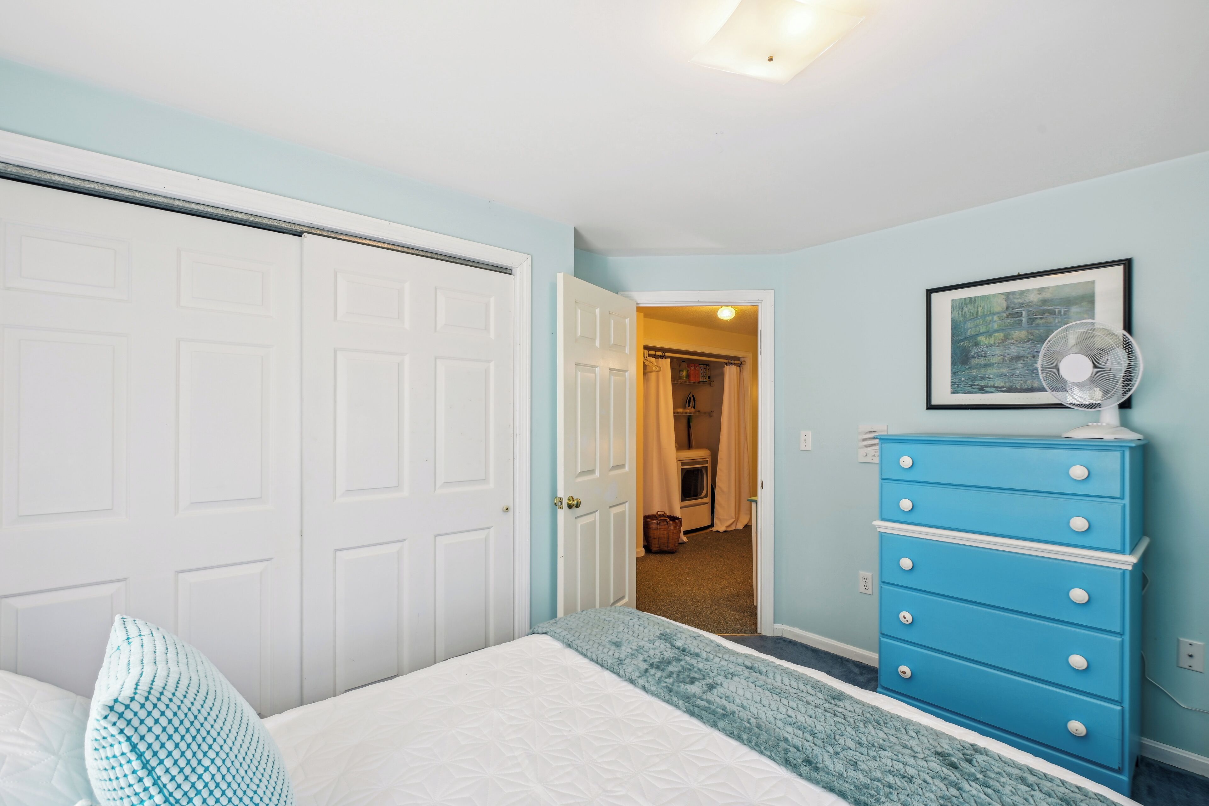 A bedroom with a bed, a blue dresser, a white door, and a hallway leading to a bathroom.
