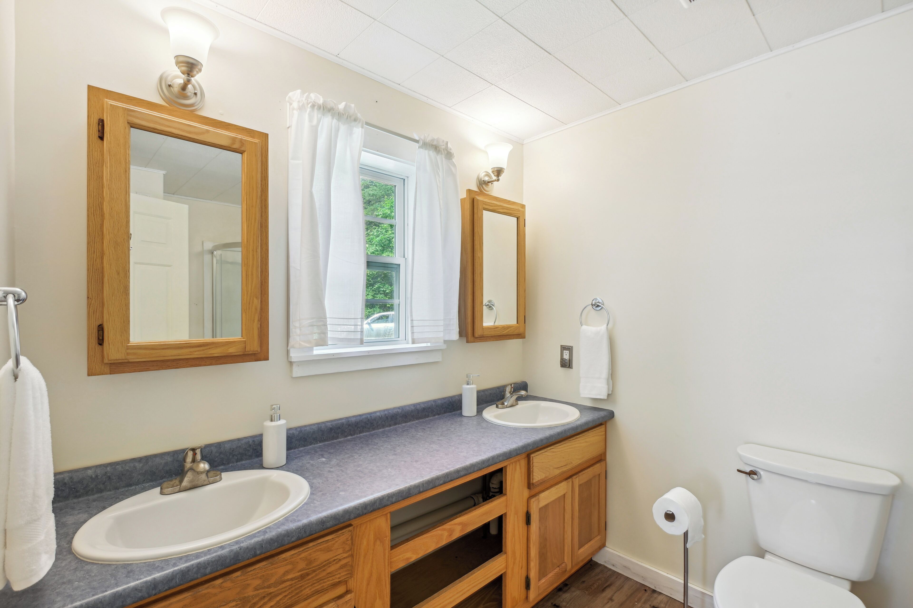 A bathroom with two sinks, wooden cabinets, a toilet, and a window with curtains.