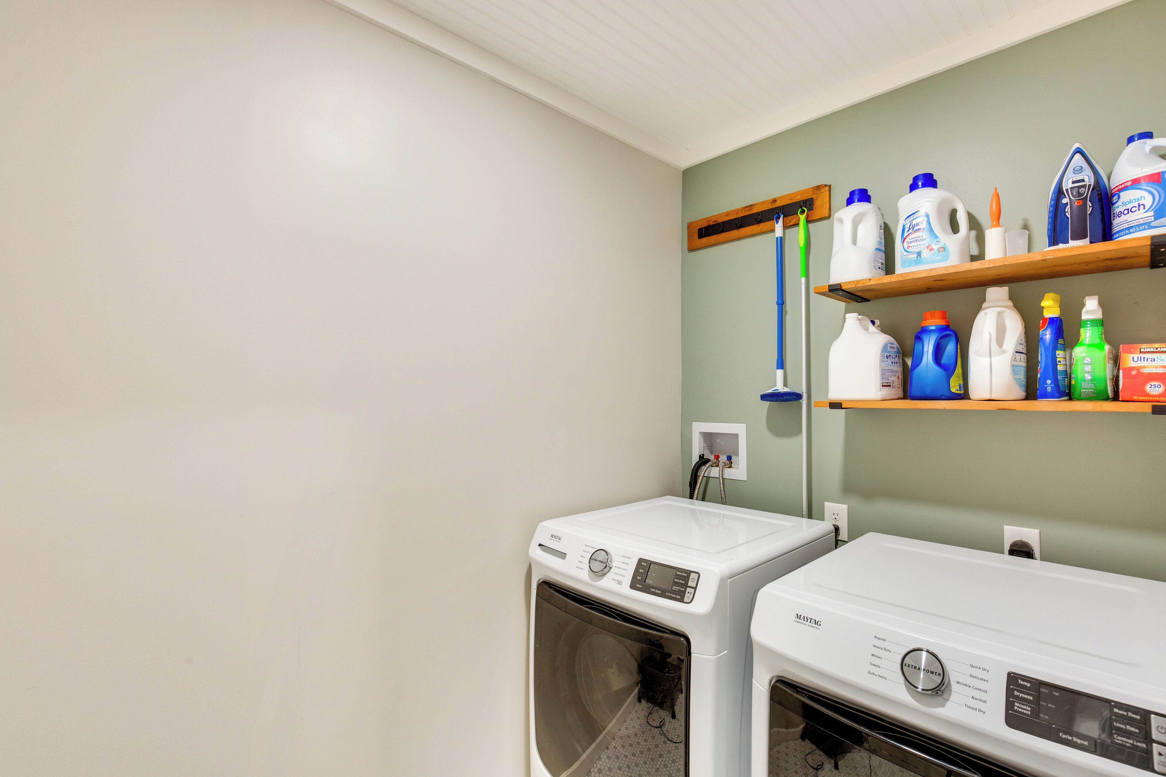 A laundry area with two front-loading washing machines, a shelf with various cleaning products, and a wall-mounted ironing board.