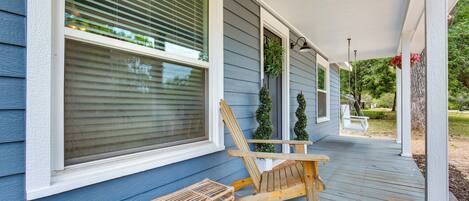 A porch with a wooden chair and a bench, a window with blinds, and a hanging planter with flowers.