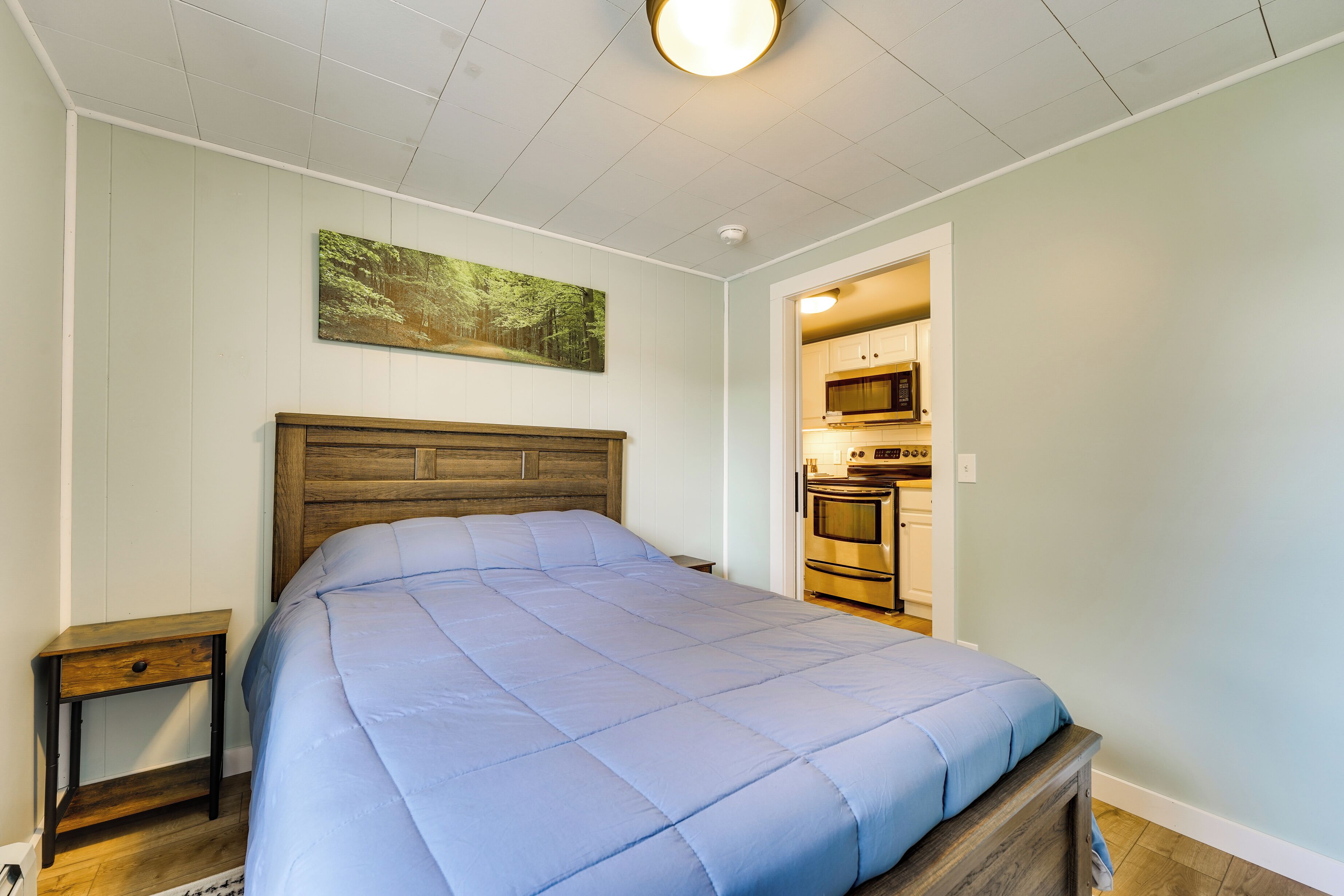 A bedroom with a wooden headboard, a blue bedspread, a nightstand, and a painting on the wall.