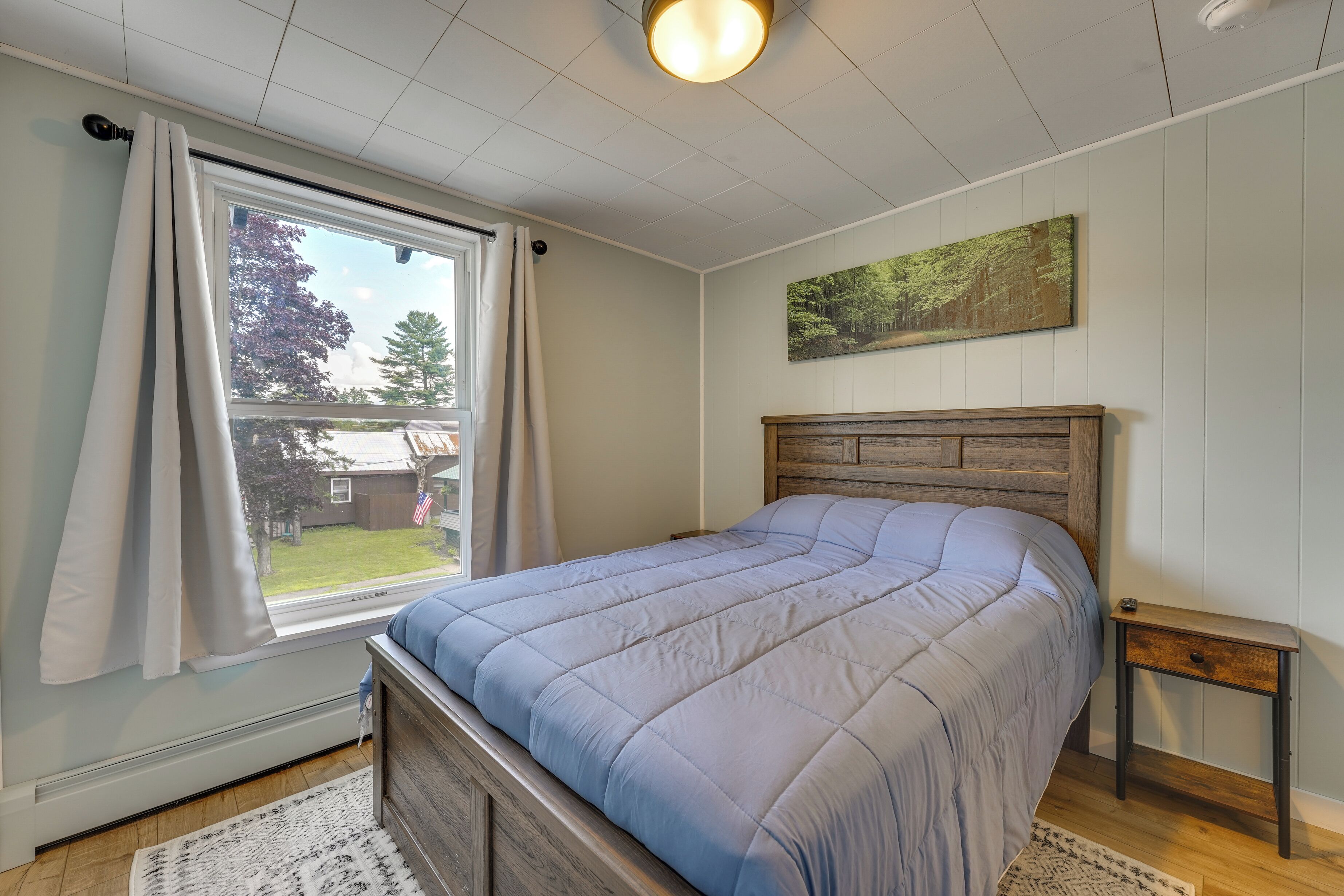 Een slaapkamer met een bed, een raam met gordijnen, een klein houten nachtkastje en een schilderij aan de muur.