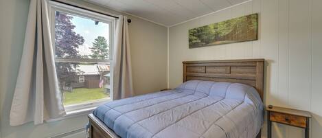 Een slaapkamer met een bed, een raam met gordijnen, een klein houten nachtkastje en een schilderij aan de muur.