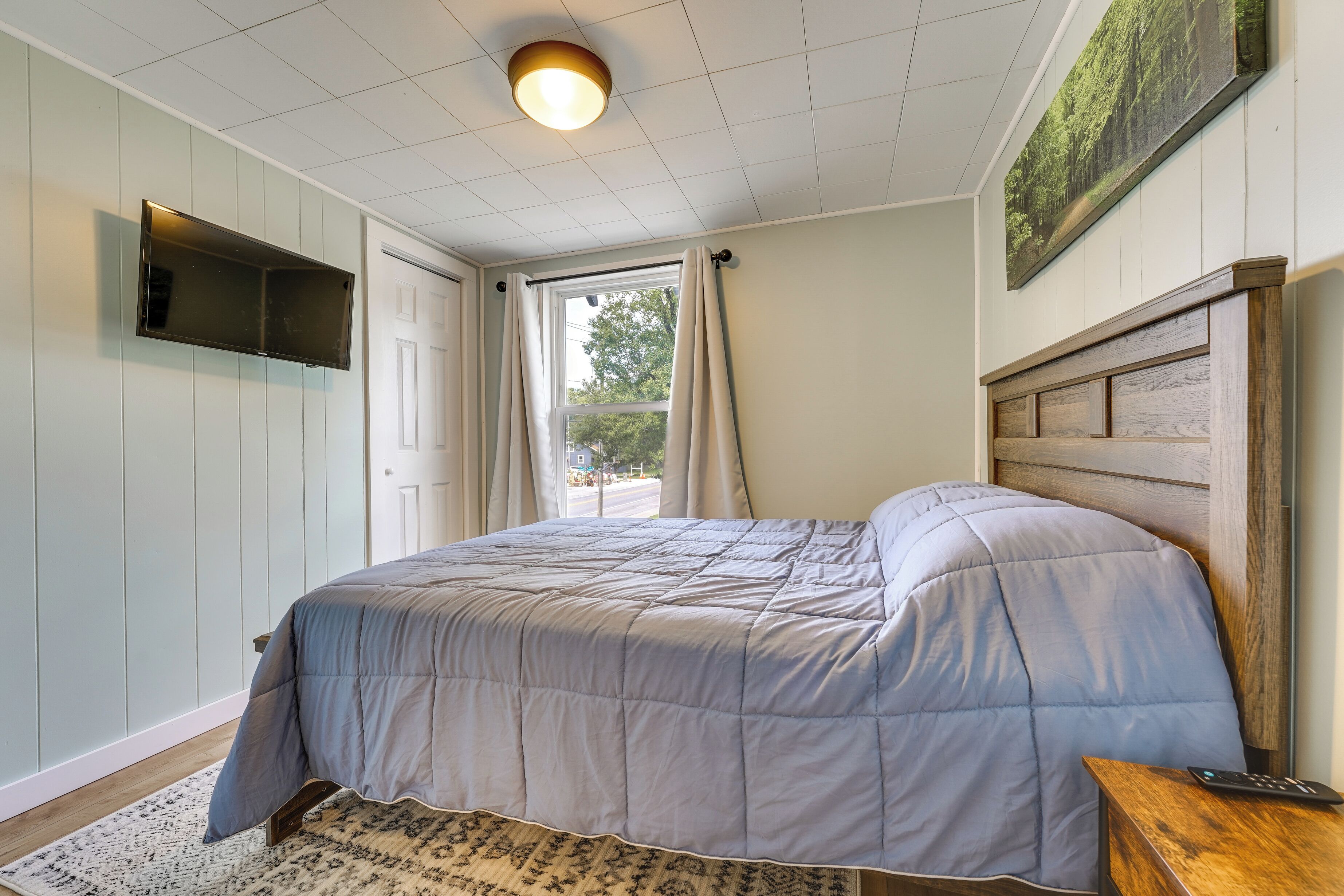 Ein Schlafzimmer mit einem Bett, einem flachbildfernseher an der Wand, einem Fenster mit Vorhängen und einem Bild über dem Bett.