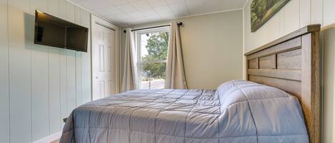A bedroom with a bed, a flat-screen TV mounted on the wall, a window with curtains, and a painting above the bed.