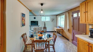 A dining area with a wooden table set for four, surrounded by chairs. A television is mounted on the wall, and there is a window with curtains. - Cozy Becket Cottage Escape w/ Lake Access! (Becket)