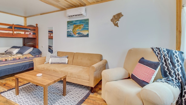 A cozy room with a wooden ceiling, a bunk bed, a beige sofa, a patterned rug, and a painting of a fish. - Cozy Norfork Cabin: Fishing, Kayaking & More! (Norfork)