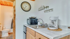 Een keuken met een magnetron, koffiezetapparaat en een spoelbak. Er hangt een klok aan de muur en een plank met potten.