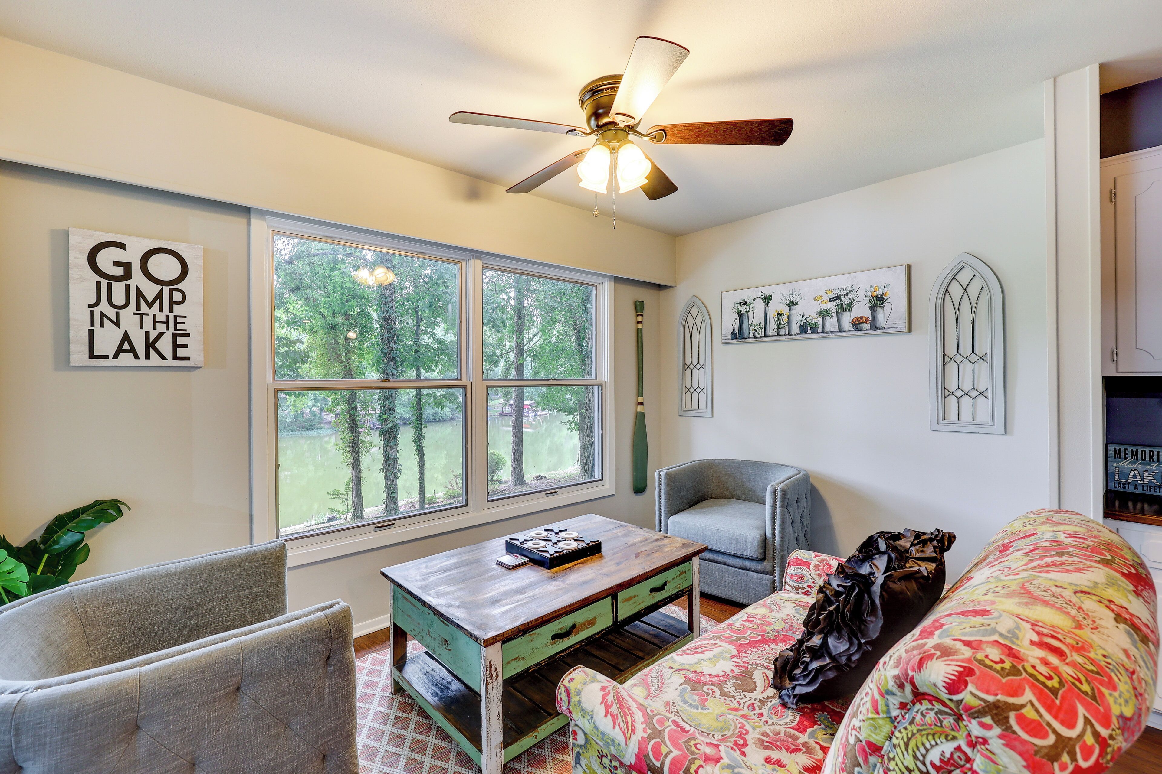 A living room with a large window, a ceiling fan, a green and white coffee table, a gray armchair, a colorful sofa, and a wall with a 'GO JUMP IN THE LAKE' sign.