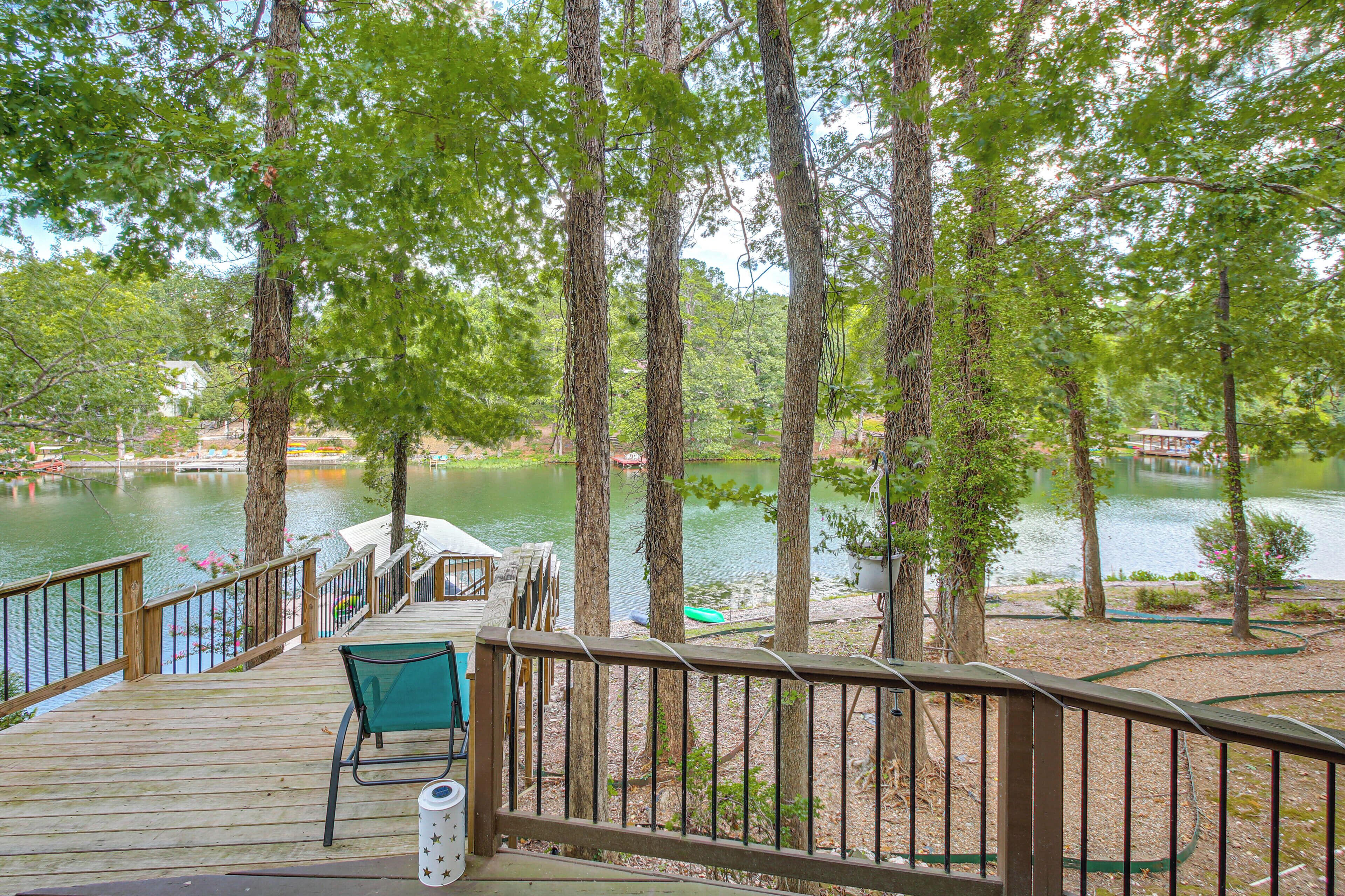 A wooden deck with a chair and a trash can, overlooking a calm lake with trees and a distant building.