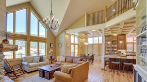 A spacious living area with a high ceiling, large windows, a chandelier, and a kitchen with bar stools. - Hot Tub + Golf Course Views: Luxe Sturgis Home! (Sturgis)