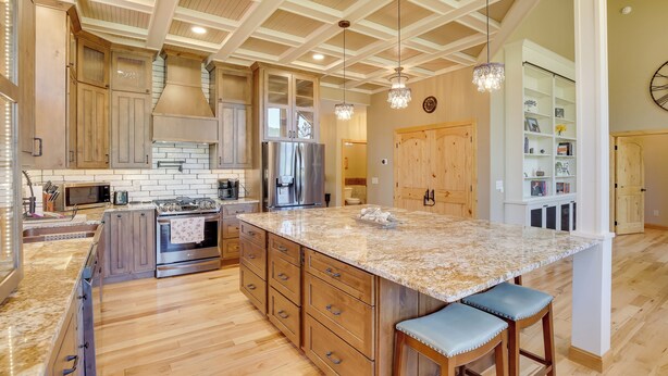 A spacious kitchen with wooden cabinets, a large island with bar stools, stainless steel appliances, and a high ceiling with exposed beams. - Hot Tub + Golf Course Views: Luxe Sturgis Home! (Sturgis)